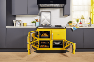 A bright yellow rayburn sits in a modern kitchen with it's doors open and cooked food inside. It is surrounded by matte grey kitchen cabinetry on a wooden floor.