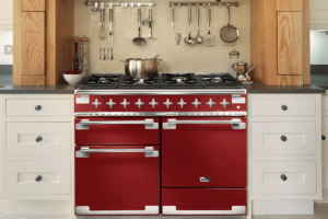 A Cherry red Falcon Elise cooker sits in kitchen surrounded by white kitchen cabinetry. Cooking utensils hang on the wall above the cooker.