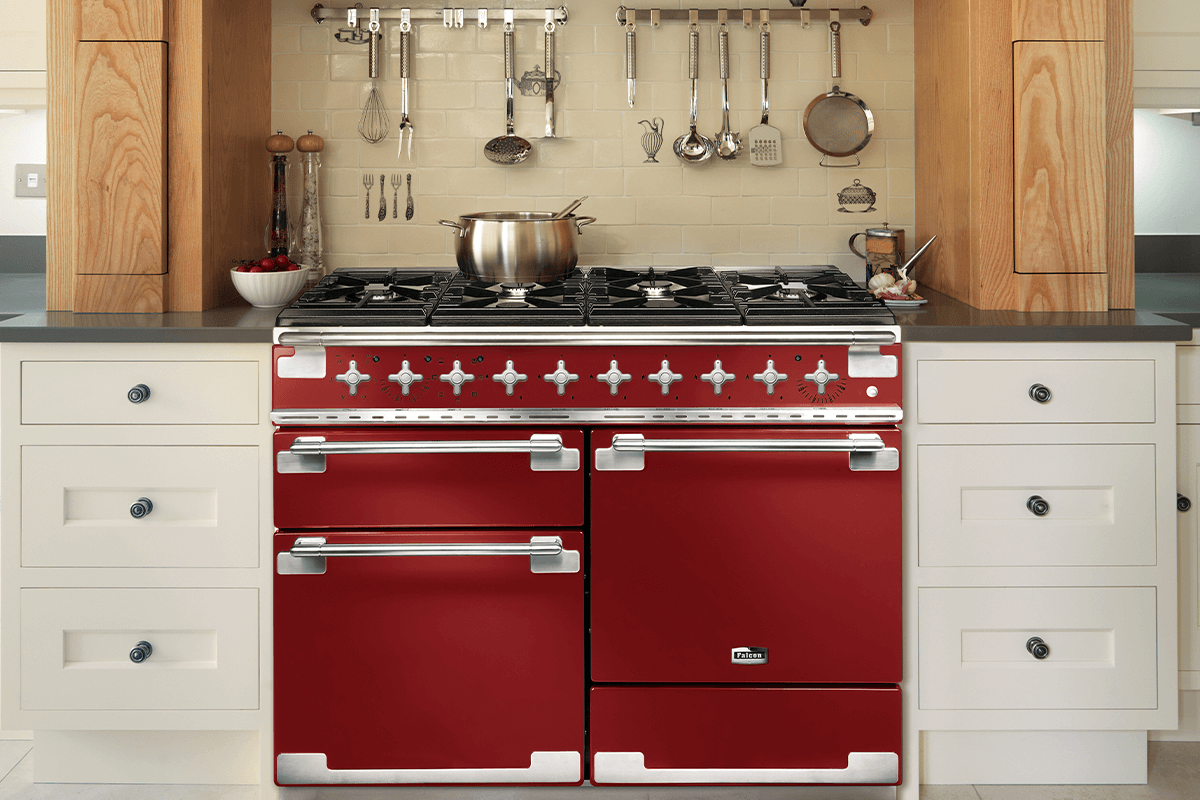 A Cherry red Falcon Elise cooker sits in kitchen surrounded by white kitchen cabinetry. Cooking utensils hang on the wall above the cooker. 