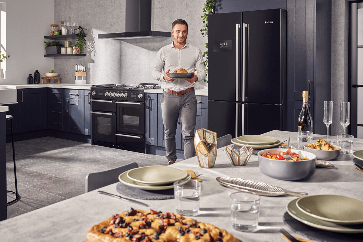 A man is walking towards a table covered in food, holding cooking in a tray. He is i a modern kitchen with grey floor tiles, blue/grey cabinets and a black Nexus steam oven. 
