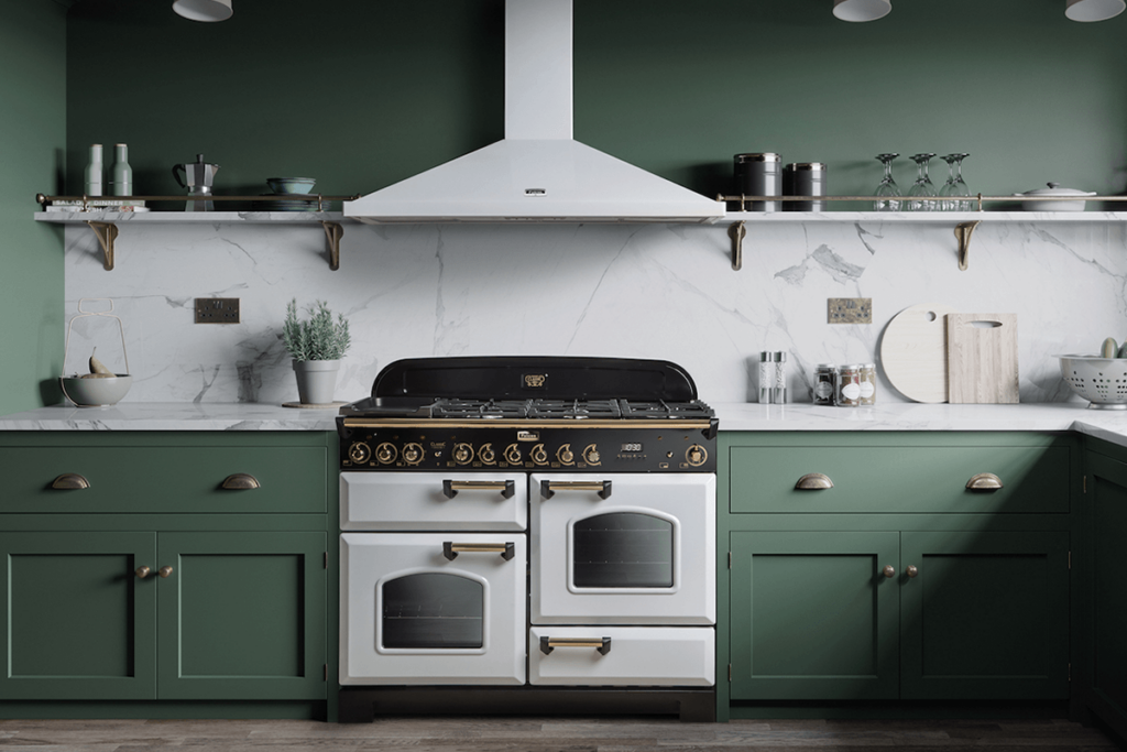 A white Falcon Classic cooker with black and gold detail with a white rangehood sits in the centre of a kitchen with olive green cabinetry, marble splashback and matching green walls with shelves above.