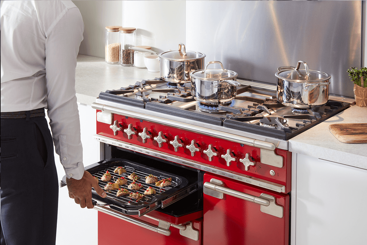 A man stands in front of a bright red Falcon Elise oven, he is removing some small grilled appetisers from the oven. Pots sit on top so the stove cooking.