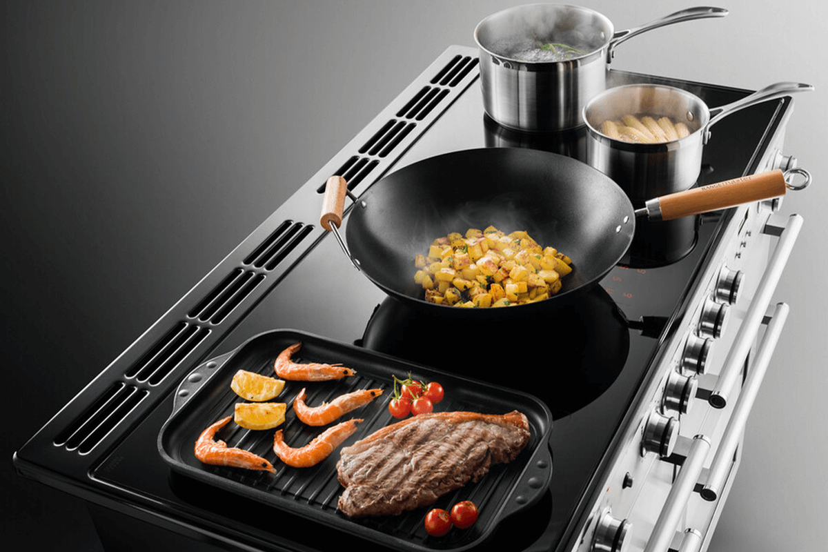 A steak, tomatoes and prawns are cooking in a griddle pan. A wok is cooking some chicken in the middle of the induction oven topo while at the back, two pots are boiling with food in them. 
