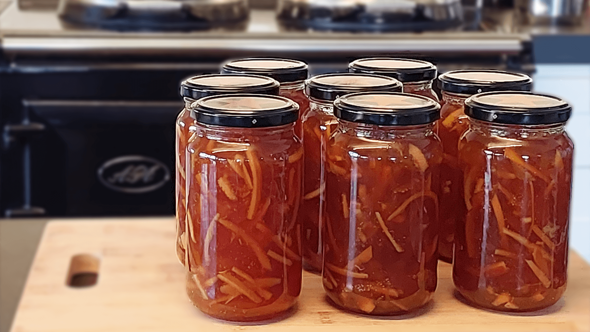 Eight jars of orange marmalade sit on top of a wooden chopping board with a dark blue AGA stove in the backround.