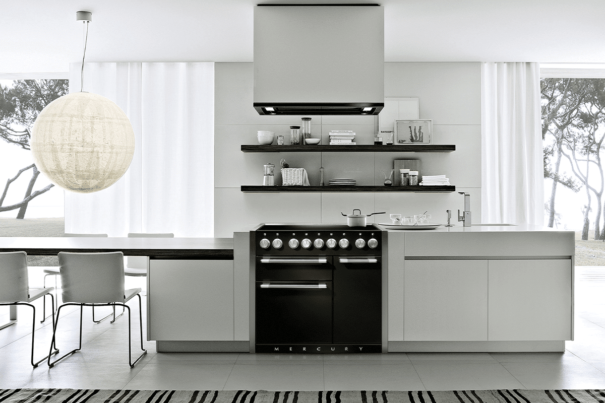 A black and chrome Falcon Nexus 90 stove sits in a modern kitchen with white cabinetry. A dining table and chairs sits to the left with a low, large hanging round light above the table. There are white curtains behind with large windows showing trees beyond.