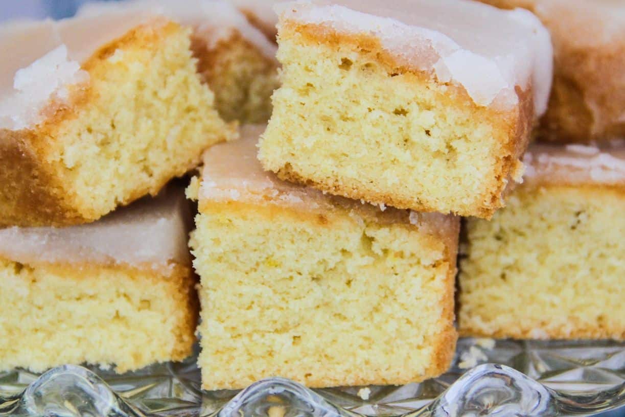 Squares of yellow golden cake with white icing are stacked on top of each other on a glass plate.
