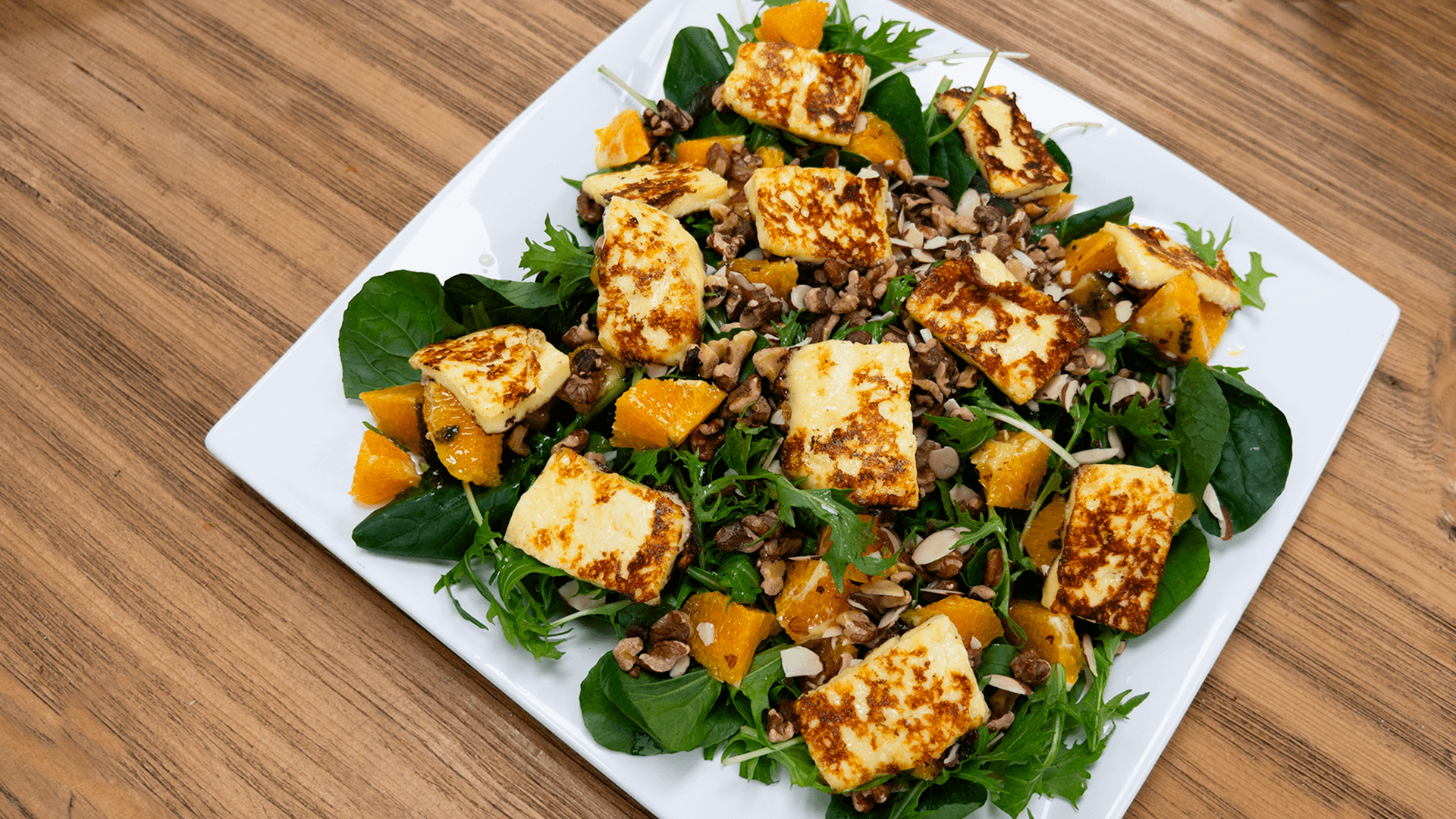 A white square plate sits on a wooden table, heaped with a Halloumi, rocket and orange salad covered in toasted walnuts.