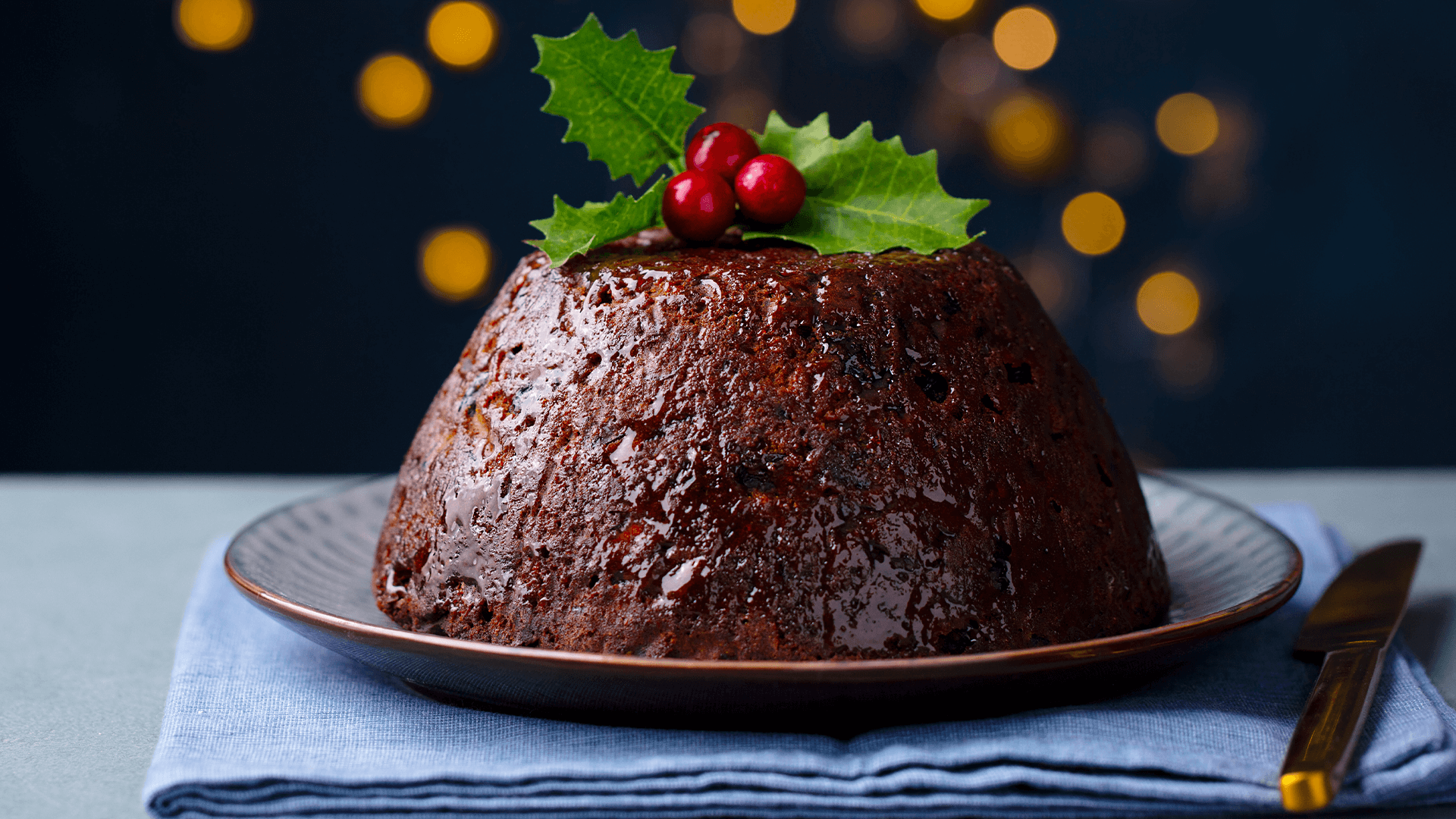 A christmas pudding sits on a dark plate on top of a blue folded napkin. Some holly foliage sits on top of the pudding, with bokeh effect lights behind on a black background.