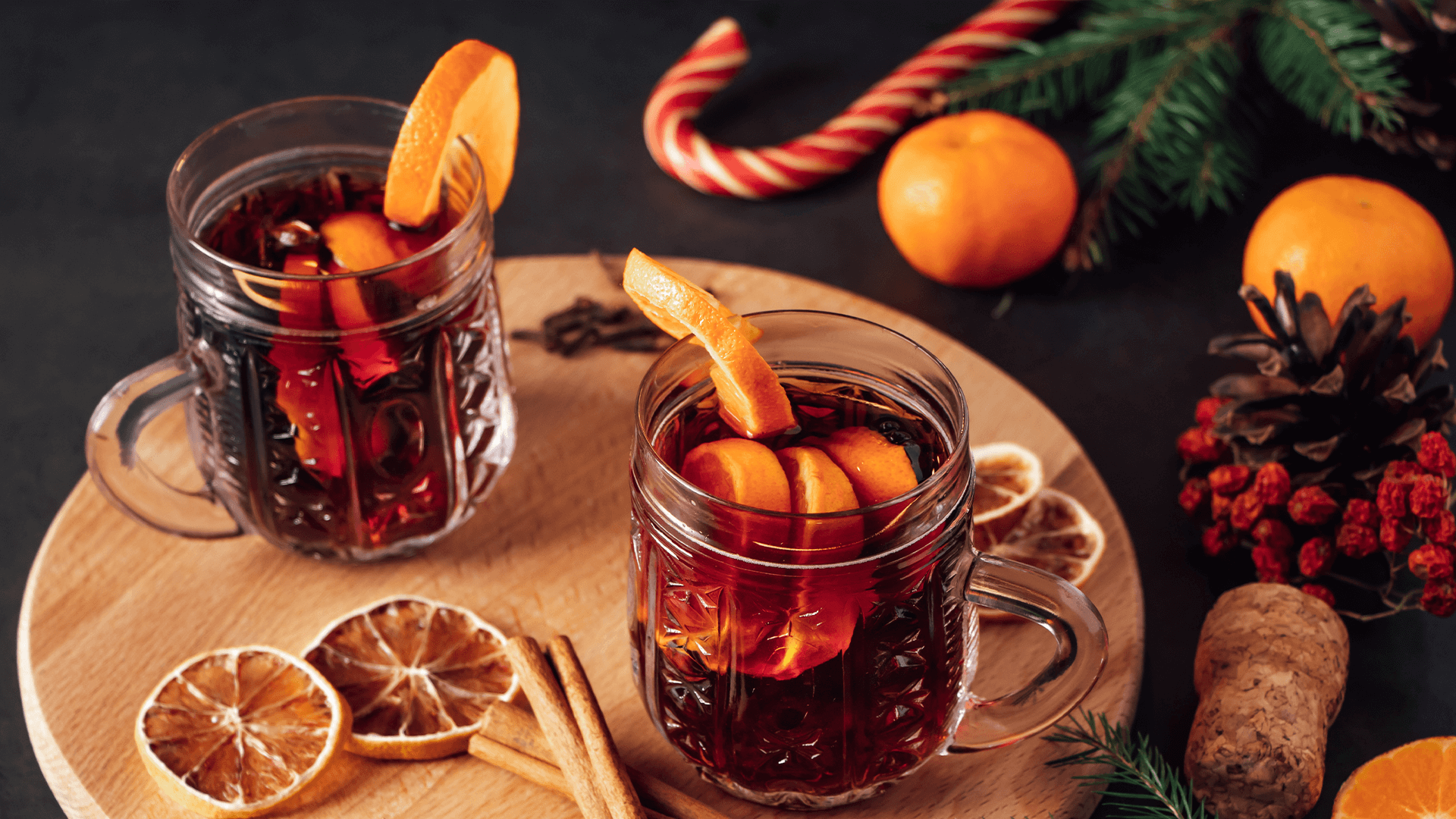 Two glasses of red mulled wine with slices of orange on the side, sit on a round wooden board with dried orange, candy canes and cinnamon sticks. In the background is pine foliage, whole oranges a cork and candy canes.