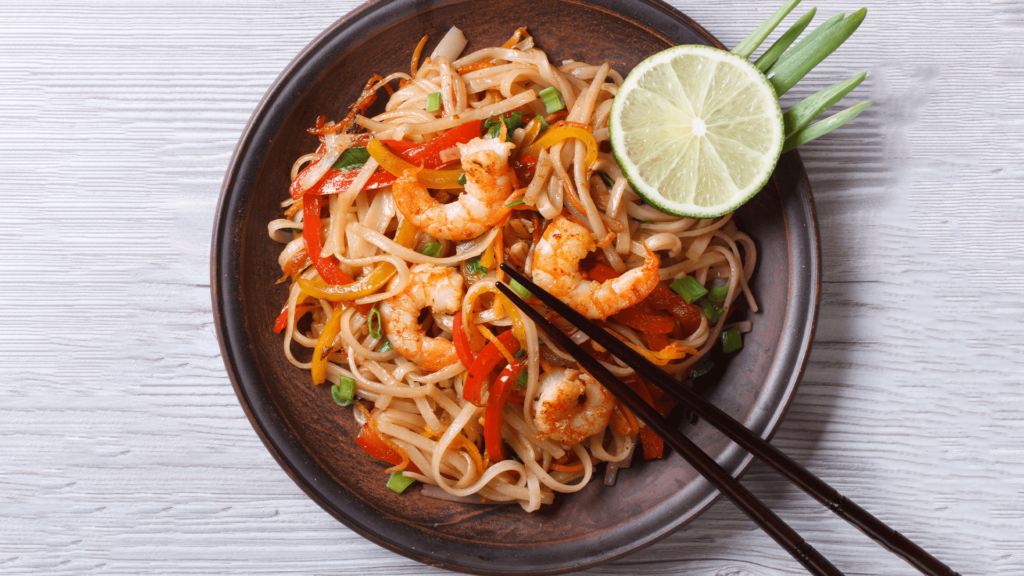 A bowl filled with noodles, prawns and vegetables, garnishes with a slice of lime and green onions with black chopsticks.