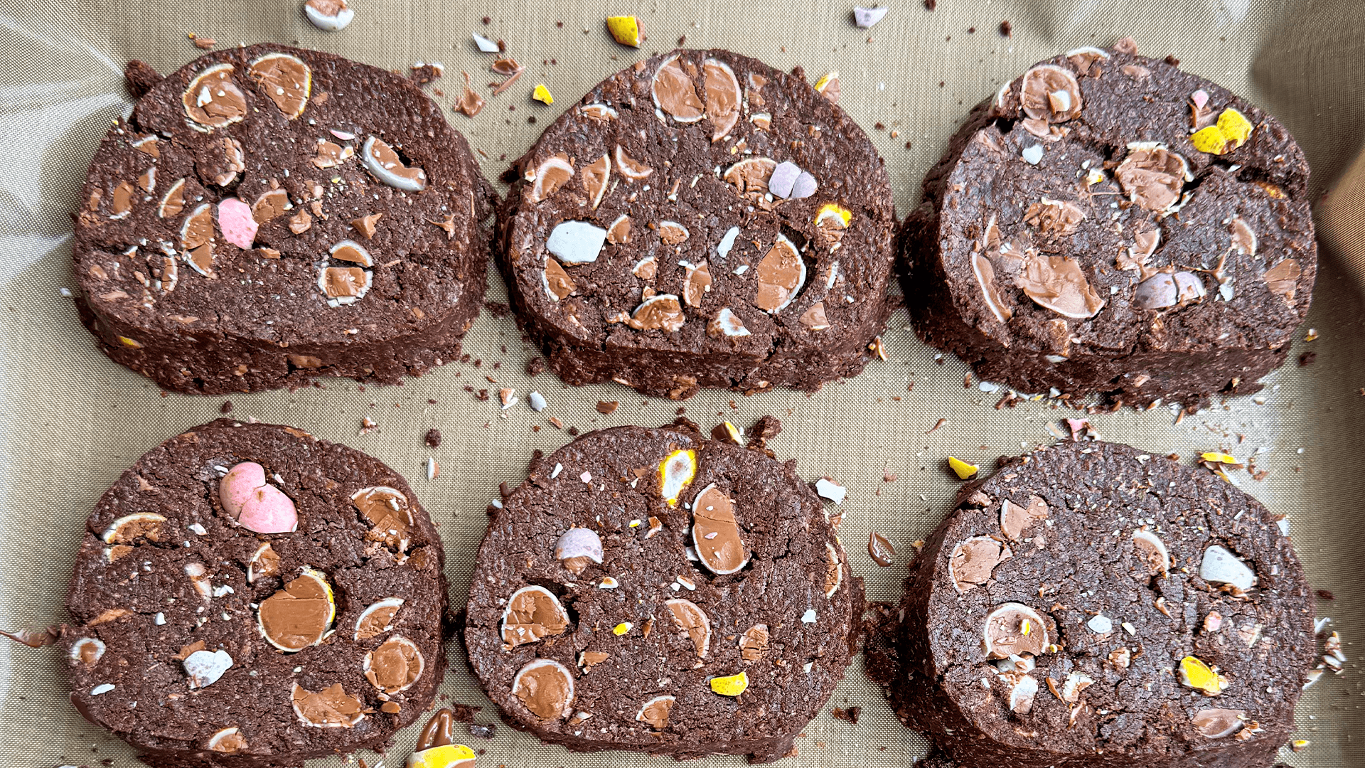 Six baked chocolate mini egg rye cookies sit on some baking paper.