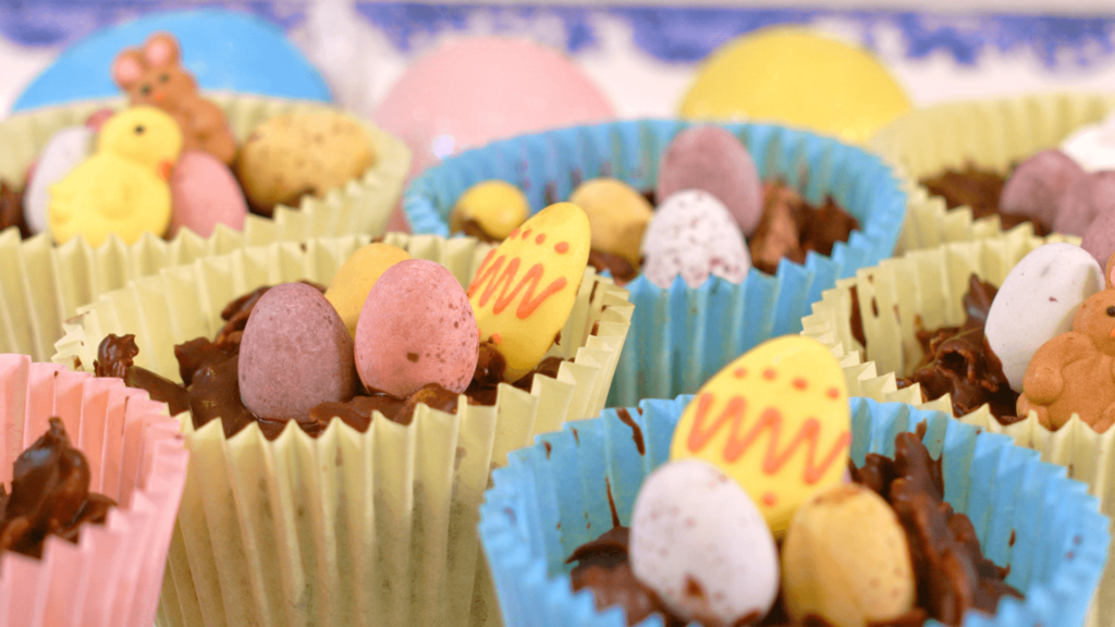 Colourful blue, yellow and pink cupcake cases filled with chocolate, mini easter eggs and cornflakes sit on a tray.