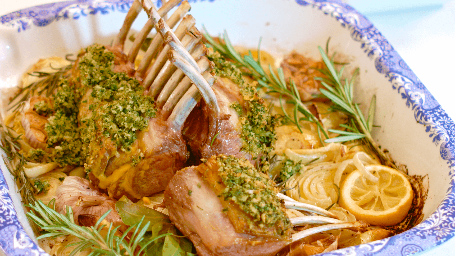 Two interlaced herb crusted lamb racks sit on a pile of roasted onions, herbs and garlic in a blue and white roasting dish.