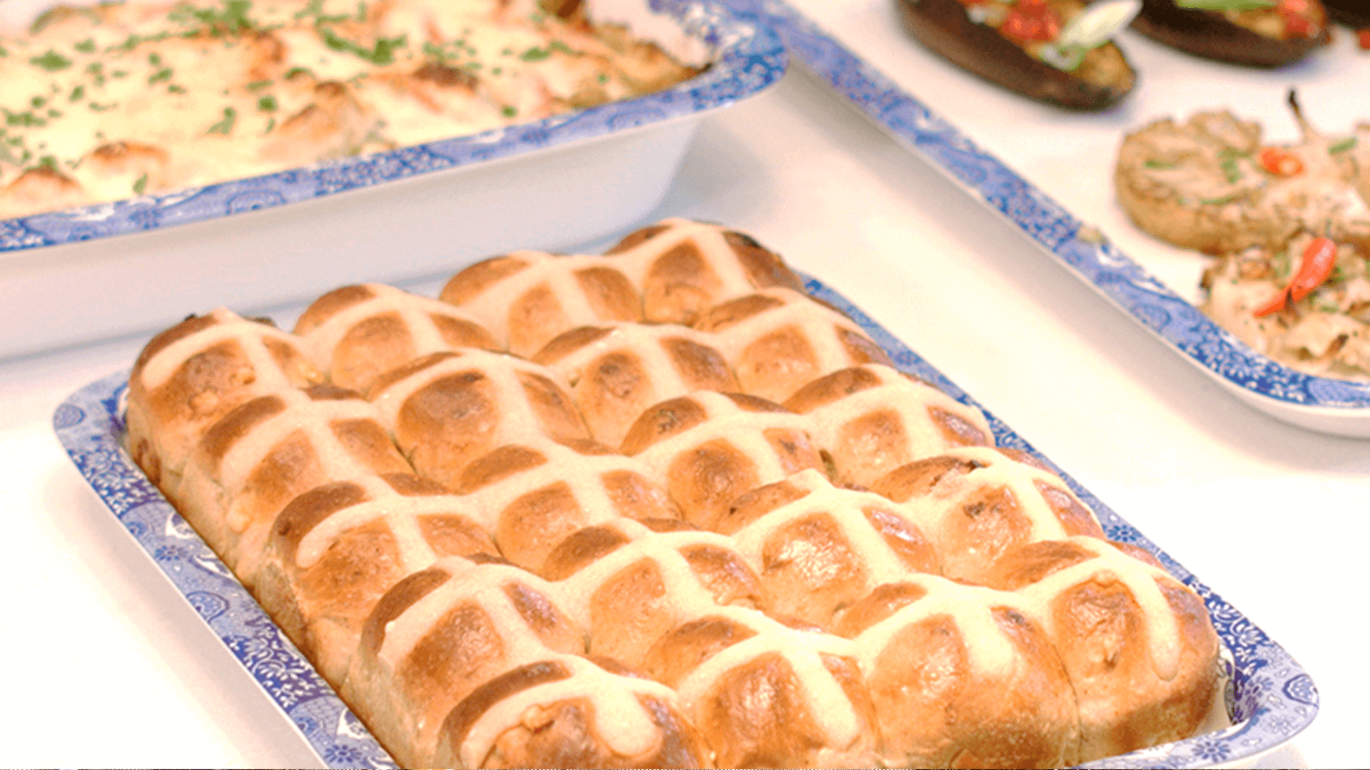 A tray of golden hot cross buns sit in a blue and white tray on a bench top.