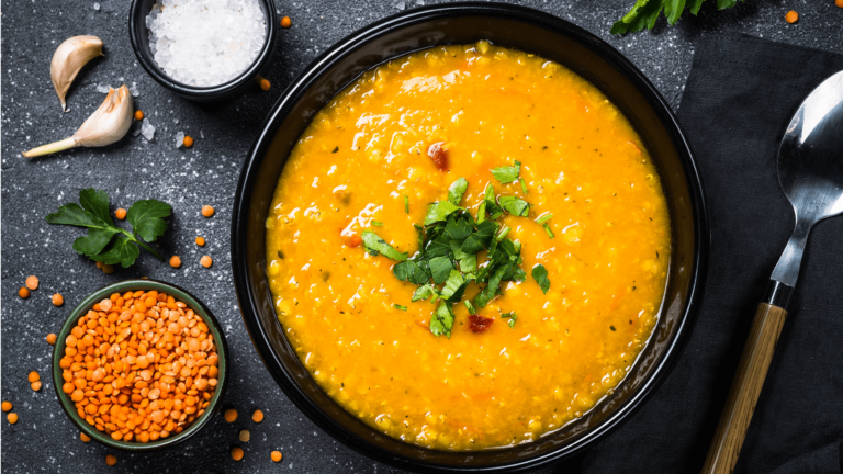 An overhead view of a bowl of carrot and lentil soup sitting inbetween a spoon and small bowl of dried red lentils, some garlic and herbs. All are sitting on a black slate board.