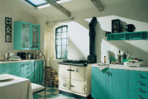 A cream Rayburn sits in an older style cottage kitchen with bright turquoise cabinetry and white and turquoise checkered lino on floor.