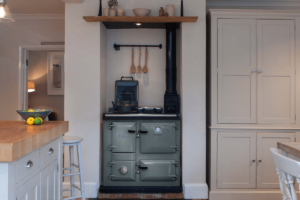 A green Rayburn sits in a white kitchen, in an old fireplace nook. A shelf is above with kitchen items and behind the cooker hang wooden cooking utensils.