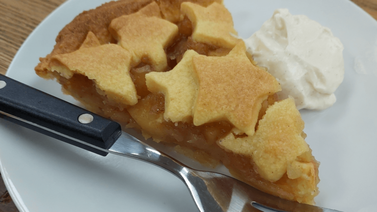 An apple pie with cut out stars crust, sits on a white plate next to some thick cream and a fork.