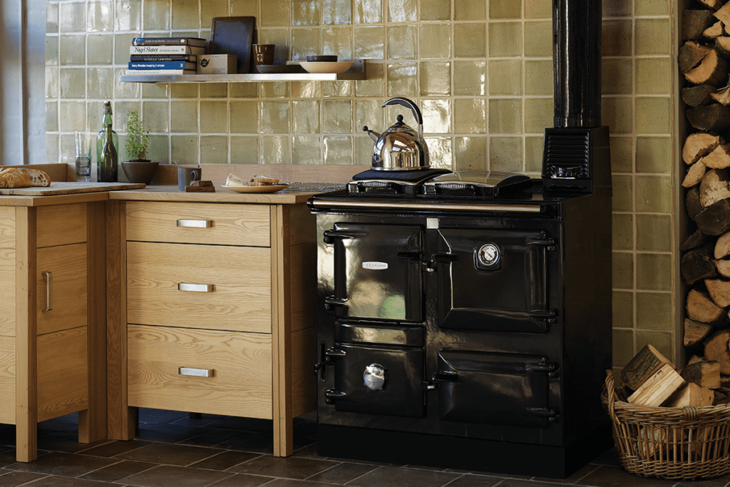 A black Rayburn cooker sits in a kitchen next to wooden cabinetry with brown green tiles on the wall. A kettle is warming on top, and piled wood sits next to the cooker.