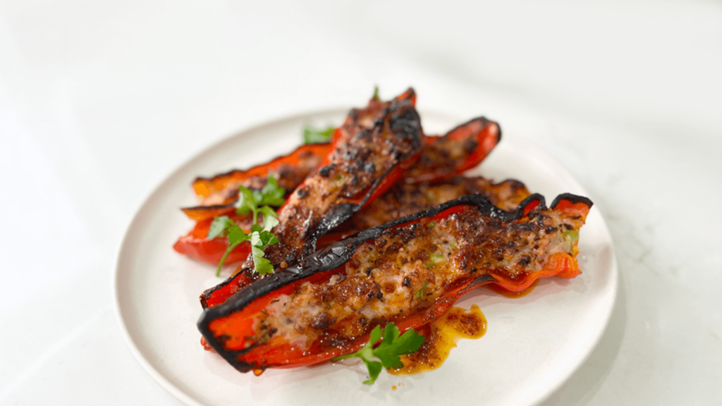 A white plate with stacked grilled red peppers stuffed with minced prawns and hers with coriander garnish sits on a marble bench top.