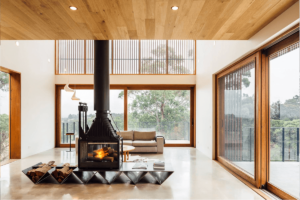 A four sided Cheminées Phillipe firebox sits in the middle of a big open planned living space with floor to celing windows all around and a wooden internal roof.