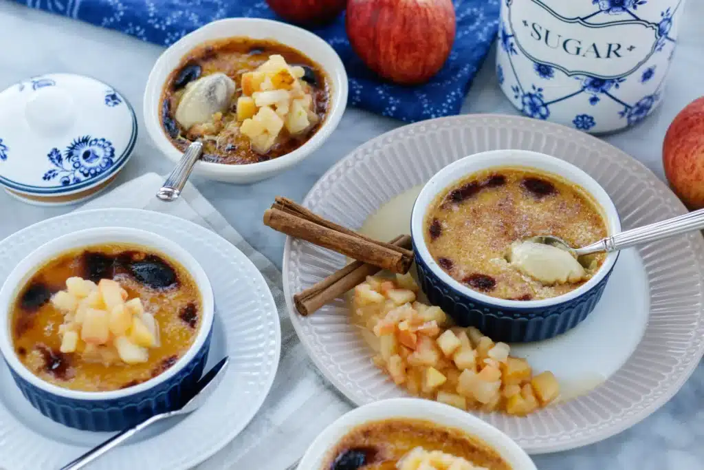 Four bowls of creme brulee topped with apple compote sit in blue bowls on white plates at a table.