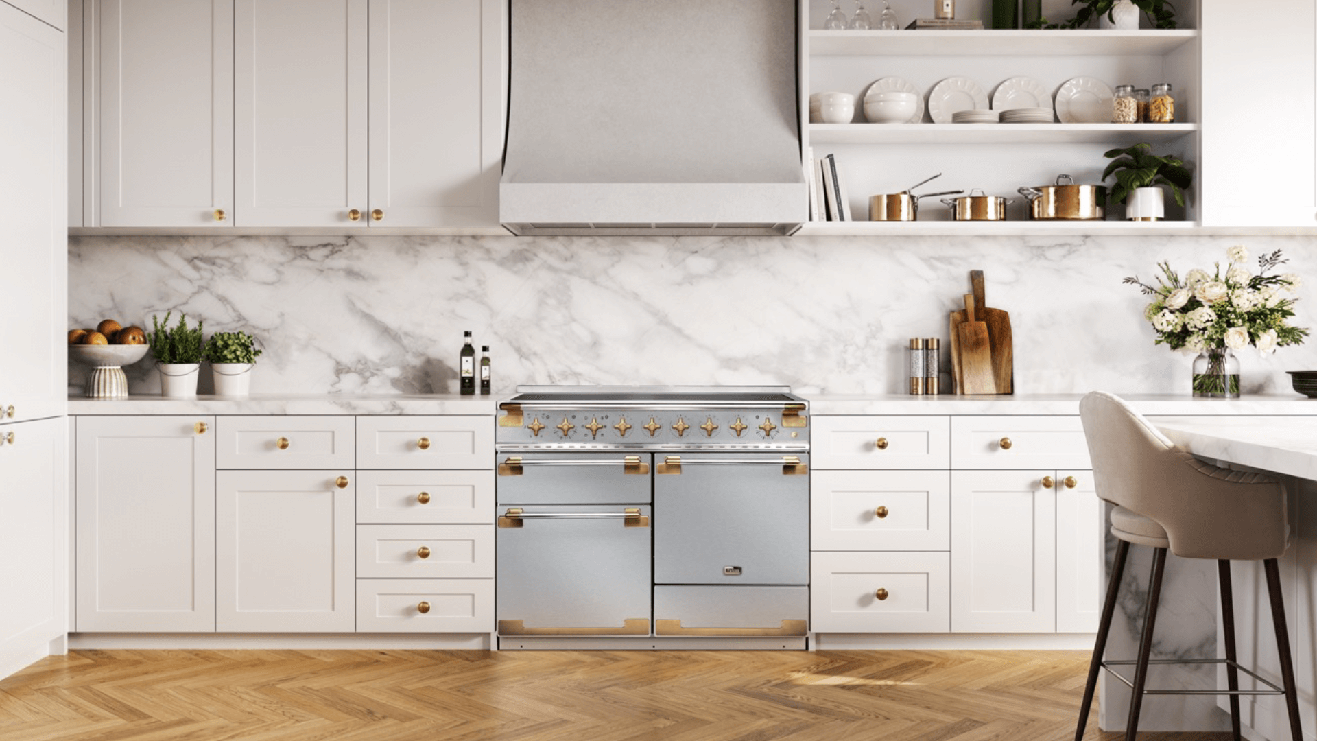A Falcon Elise in stainless steel with chrome and brass detail sits in modern kitchen with white cabinetry, parquet floowring and a bench with bar stools in the foreground. There are breadboards, fruit bowls and vased flowers on the bench either side of the cooer and a large built in rangehood above the cooker.