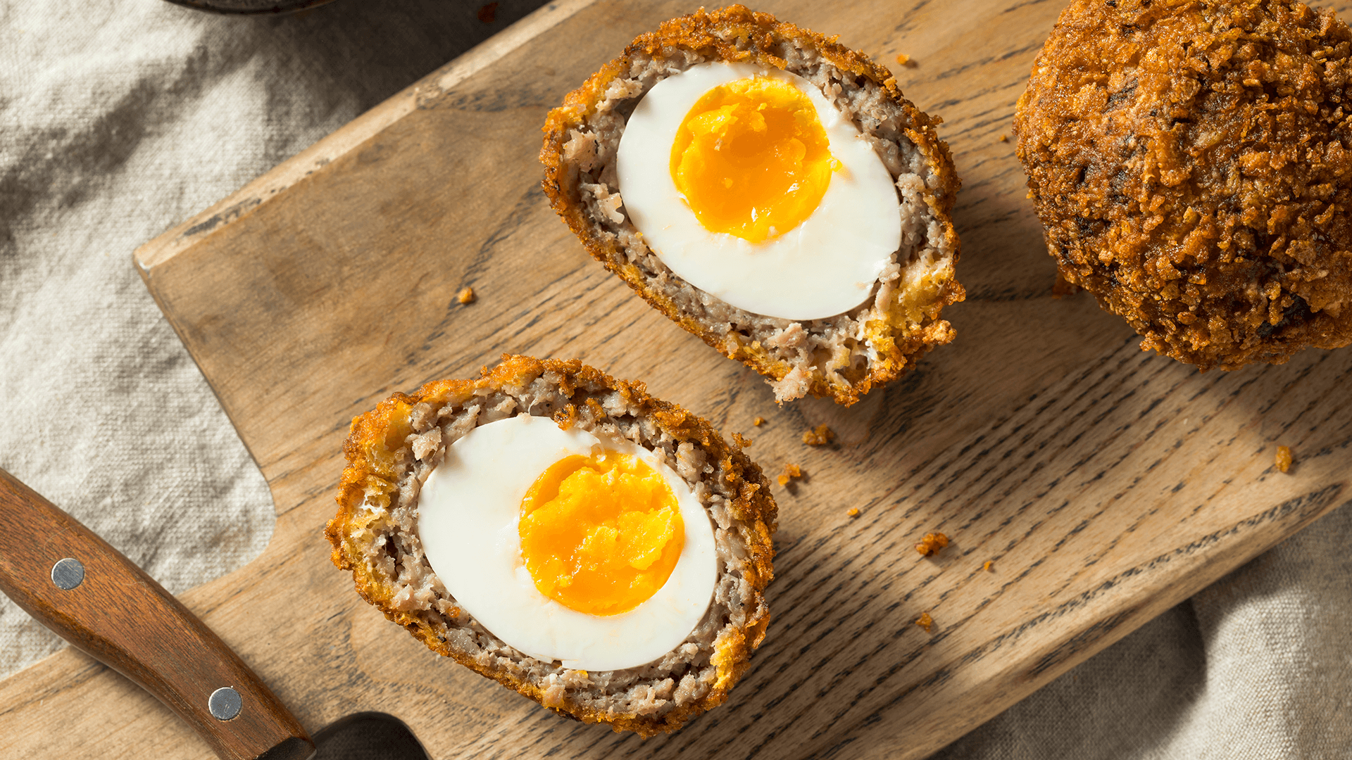 An overhead view of a scotch egg sliced in half, sitting on a wooden board with a knife.