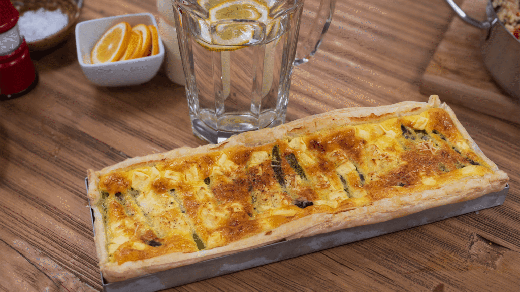 A silver baking tray of cooked asparagus tart, with golden melted cheese on top, sits on a wooden kitchen table, with a glass and a dish of cut up lemons in the background.