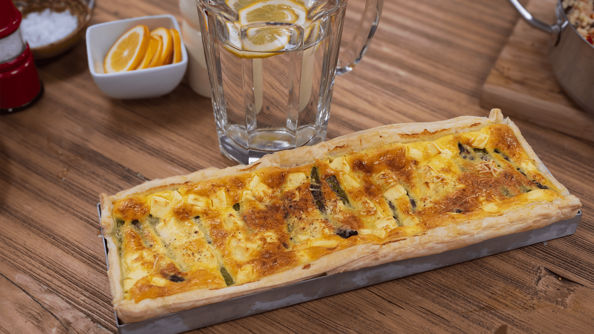 A silver baking tray of cooked asparagus tart, with golden melted cheese on top, sits on a wooden kitchen table, with a glass and a dish of cut up lemons in the background.